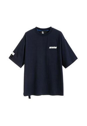 Persona con cabello rubio de espaldas, lleva una camiseta negra de gran tamaño con un gráfico de ojo y pantalones anchos con estampados florales en tonos rosa.