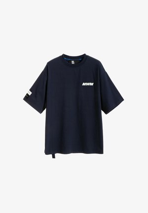 Persona con cabello rubio de espaldas, lleva una camiseta negra de gran tamaño con un gráfico de ojo y pantalones anchos con estampados florales en tonos rosa.