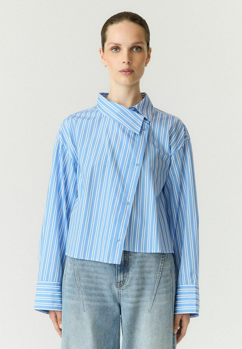 Femme aux cheveux attachés portant une chemise à rayures verticales bleues et blanches avec un col asymétrique et un jean bleu clair.