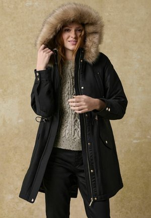 Parka noire avec une capuche en fourrure, fermeture éclair et poches latérales. Portée par-dessus un pull en tricot torsadé gris et un pantalon noir. Texture lisse.