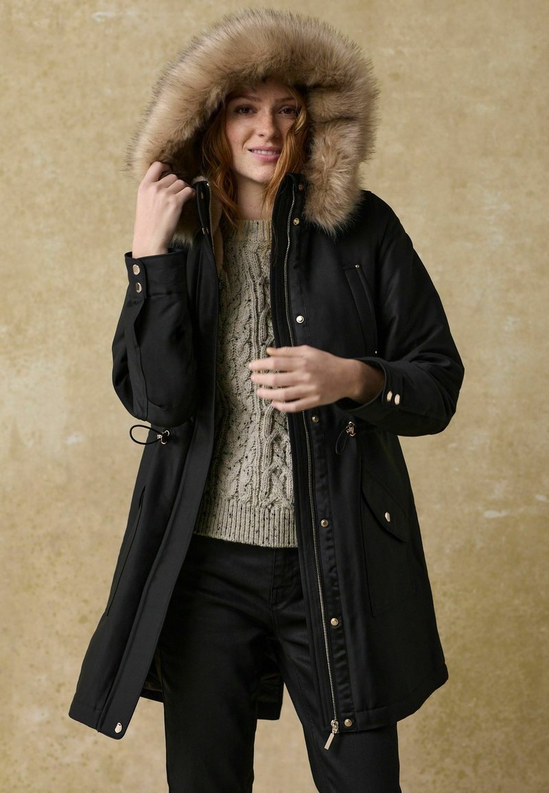 Parka noire avec une capuche en fourrure, fermeture éclair et poches latérales. Portée par-dessus un pull en tricot torsadé gris et un pantalon noir. Texture lisse.