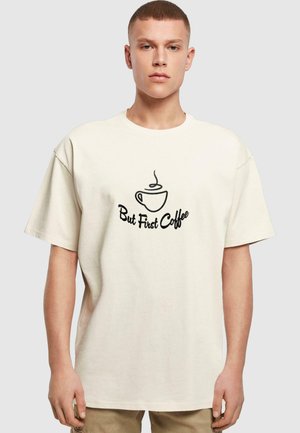 Cremefarvet oversized t-shirt med en sort grafik af en kaffekop med damp og teksten "Men først kaffe" i en buet skrifttype.