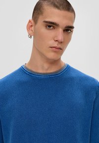 Blauer strukturierter Strickpullover mit Rundhalsausschnitt, über einem grauen Shirt getragen, mit langen Ärmeln und einer lockeren Passform.