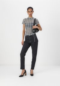 Sheer, patterned top in shades of gray, paired with high-waisted black jeans and black heels with a chunky heel. Holds a small black bag.