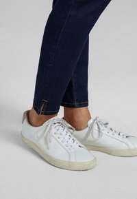 Zapatillas de cuero blancas con punta redonda, suela plana de goma y cierre con cordones. Combinadas con jeans de color azul oscuro con un dobladillo cosido en color naranja.