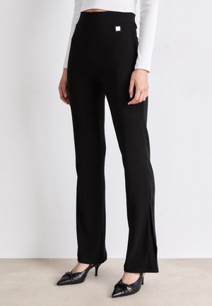 Mujer que lleva pantalones negros de talle alto y corte recto con aberturas laterales, camiseta blanca de manga larga y tacones negros puntiagudos con detalle de lazo.
