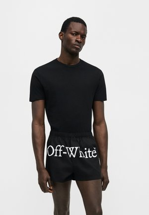 Mężczyzna noszący czarną koszulkę z krótkim rękawem i czarne krótkie spodenki z napisem "Off-White" dużymi białymi literami na przednim pasie.