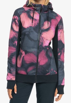 Femme portant un sweat à capuche zippé noir et rose avec motif floral et des trous pour les pouces, debout devant un fond blanc.