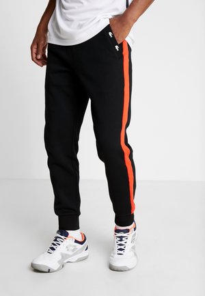 Man die zwarte joggingbroeken draagt met rode strepen aan de zijkant, witte sneakers met marineblauwe en oranje accenten, terwijl hij met één hand in zijn zak staat.