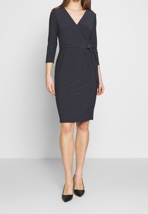 Femme portant une robe portefeuille noire longueur genou à pois blancs, manches trois-quarts, associée à des escarpins noirs à bout pointu.