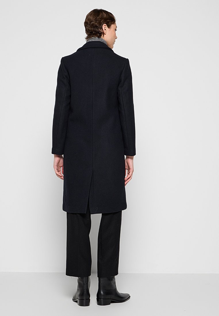 Personne se tenant de dos, portant un long manteau noir, un pantalon noir, des bottines noires et un pull col roulé gris.