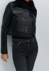 Czarna skórzana kurtka typu cropped z kołnierzem z sztucznego futra, z przednimi kieszeniami, zapięciem na guziki i gładką teksturą. Noszona z czarnymi dżinsami.