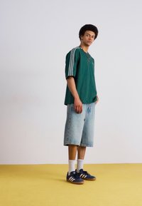 Grøn oversized t-shirt med tre hvide striber, lyseblå denimshorts, marineblå sneakers med hvide accenter og hvide sokker.