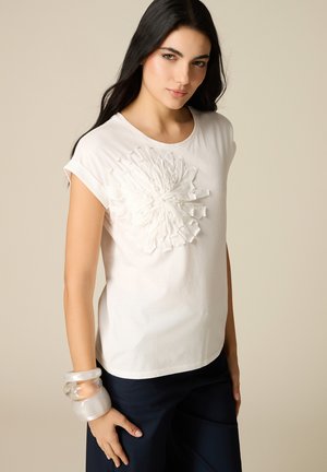 Donna con capelli lunghi e scuri che indossa una maglietta bianca a maniche corte con dettaglio floreale strutturato e pantaloni scuri, accessoriata con grandi braccialetti trasparenti.