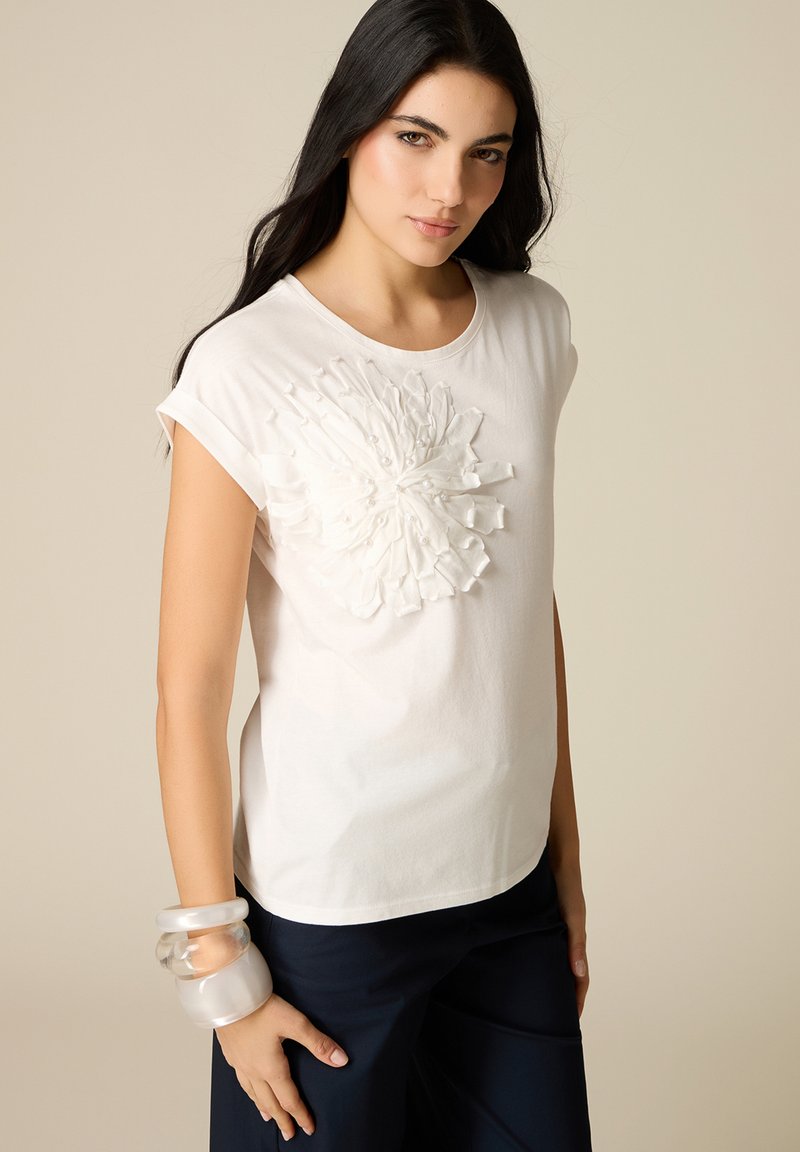 Mujer con cabello largo y oscuro, vistiendo una camiseta blanca de manga corta con detalle floral texturizado y pantalones oscuros, complementada con grandes pulseras transparentes.