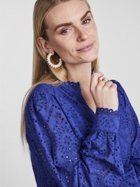 Kungablå ögla-blus med intrikata broderade mönster och scalloped trim, tillsammans med stora pärl-hängande örhängen. Slät textur.