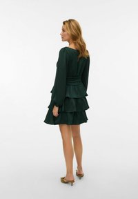 Zaļas krāsas daudzslāņu kleita ar garām piedurknēm, savāktu jostasvietu un volāniem apakšmalā. Gluda auduma tekstūra un izteikta silueta forma. Izskatu papildina brūnas krāsas augstpapēžu apavi.