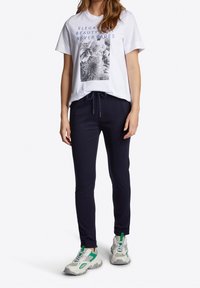 Vit t-shirt med blommig grafik och blå text, kombinerad med marinblå joggers med dragsko i midjan, samt ljusgrå sneakers med gröna detaljer.