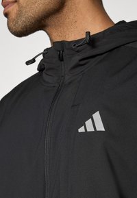 Μαύρο μπουφάν με φερμουάρ μπροστά, ρυθμιζόμενο κουκούλα και ανακλαστικό λογότυπο στο στήθος. Διαθέτει κομψή, λεία υφή και εφαρμοστή σχεδίαση.