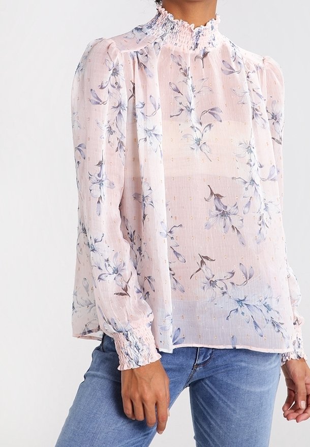 Femme porte un chemisier rose clair transparent avec un col haut volanté et des poignets, avec un imprimé floral bleu, associé à un jean bleu.