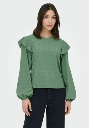 Femme portant un chemisier vert à manches longues avec broderies florales et épaules volantées, associé à un pantalon noir sur un fond uni.