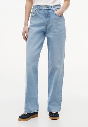 Άτομο που φοράει ανοιχτό μπλε τζιν υψηλόμεσα με ίσιο πόδι, με το χέρι στην τσέπη και μαύρα casual sneakers με καουτσούκ σόλες.