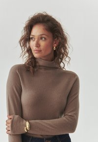 Smeđi turtleneck pulover napravljen od mekane, teksturirane tkanine. Ima duge rukave s otvorima za palčeve, uparen s zlatnom narukvicom.