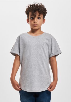 Dreng med krøllet hår iført en grå kortærmet skjorte og blå bukser, stående med fronten mod kameraet mod en ensfarvet hvid baggrund.