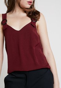 Mujer con una blusa sin mangas color burdeos con tirantes de hebilla negra y pantalones oscuros, contra un fondo claro y liso.