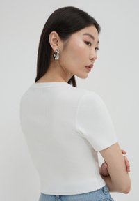 Giovane donna in un top a maniche corte bianco in maglia e jeans blu, con capelli scuri e lisci, che indossa grandi orecchini a cerchio in argento, rivolta di lato.