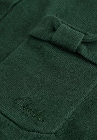 Skogsgrön textil med struktur som har en fickdetalj med sladd och subtil branding i sydda bokstäver.
