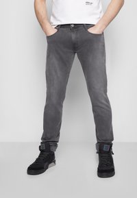 Homme portant un jean slim gris délavé et des baskets noires, debout les mains dans les poches devant un fond uni.