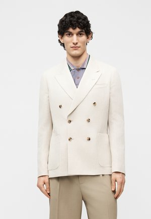 Giovane uomo con capelli ricci scuri indossa un blazer doppiopetto color crema sopra una camicia a righe con colletto e pantaloni beige, in piedi davanti a uno sfondo semplice.