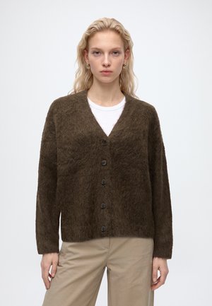 Jeune femme aux cheveux blonds portant des boucles d'oreilles en or, un cardigan marron duveteux boutonné par-dessus une chemise blanche et un pantalon beige, debout devant un fond blanc.