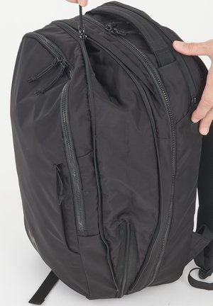 Sac à dos noir avec plusieurs compartiments zippés, l’un partiellement ouvert, tenu par deux mains sur un fond blanc.