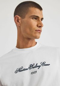 Biała bawełniana koszulka z granatowym haftowanym napisem "Nautica Sailing Crew 1983" w stylu kursywy. Klasyczny fason z okrągłym dekoltem.