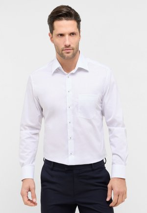 Mand med kort brunt hår iført en hvid skjorte med lange ærmer og mørke bukser, stående mod en ensartet lys baggrund.