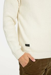 Weißer, gerippter Strickpullover mit einer strukturierten Oberfläche und einem schwarzen Etikett am Saum. Der Pullover hat eine lockere Passform und einen Rundhalsausschnitt.