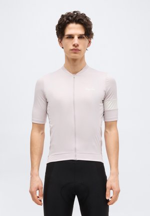 Joven con una camiseta de ciclismo de manga corta color gris claro con cremallera frontal y pantalones cortos de ciclismo acolchados negros, posando frente a un fondo liso.