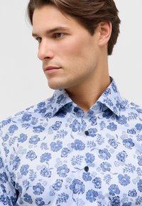 Lichtblauw bloemenoverhemd met donkerblauwe bloemenpatronen, een puntkraag en zwarte knopen. Zachte stoftextuur.