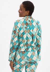Blaue und braune Karojacke mit langen Ärmeln, die akzentuiert ist durch ein Leopardenmuster und ein Patch-Label auf der Rückseite. Strukturierter Stoff.