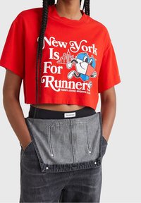 Červené zkrácené tričko s grafikou "New York je pro běžce", spárované s černými džíny s šedou kapsou a patentkami.
