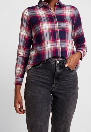 Persona con una camisa de cuadros de manga larga en tonos púrpura, rosa, gris y blanco, metida dentro de unos vaqueros negros de cintura alta, con una mano en el bolsillo.