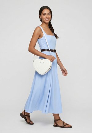 Junge Frau mit geflochtenem Haar, die ein hellblaues Kleid, braune Sandalen und eine weiße, herzförmige Umhängetasche trägt, geht lächelnd spazieren.