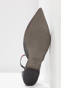 Chaussure pour femme à bout pointu avec bride de talon à motifs, semelle marquée "Lavorazione Artigianale" et taille 37, présentée debout contre un mur.