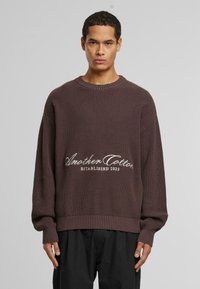 Brązowy sweter robiony na drutach z prążkowanym wzorem, z opuszczonymi ramionami, z okrągłym dekoltem oraz białym haftem z napisem "Another Cotton ESTABLISHED 2023."