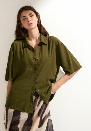 Jeune femme aux longs cheveux bruns portant une chemise oversize vert olive à boutons et une jupe à motifs, se tenant contre un mur blanc.