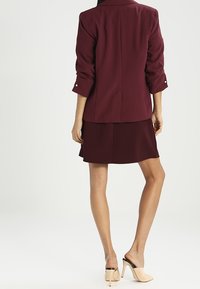 Burgundská sako s ohrnutými rukávy a knoflíkovými detaily, v kombinaci s ladící burgundskou sukní. Světle béžové sandály na otevřené špičce dotvářejí celý vzhled.