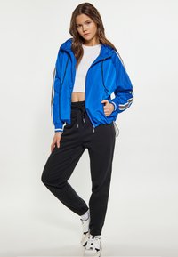 Giacca leggera blu con zip e cappuccio, dettagli bianchi sulle maniche; abbinata a pantaloni jogger neri e scarpe da ginnastica bianche.