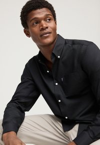 Young man wearing a black button-up shirt and beige pants, sitting and looking to his left against a plain background.